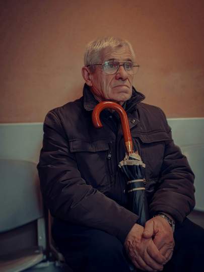 Elderly man wearing glasses and a dark coat sitting in a waiting room holding a closed umbrella with both hands, looking slightly to the side with a calm serious expression.