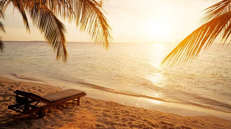Golden hour serenity a cushioned lounge chair faces the tranquil ocean as palm fronds frame a glowing tropical sunset.